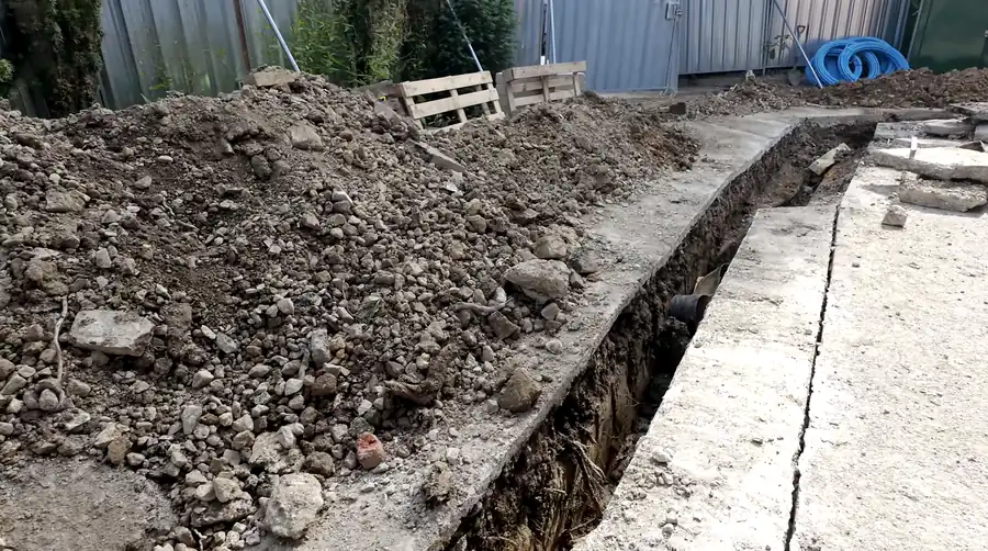 Open utility trench excavated beside a concrete path, with spoil heaped alongside