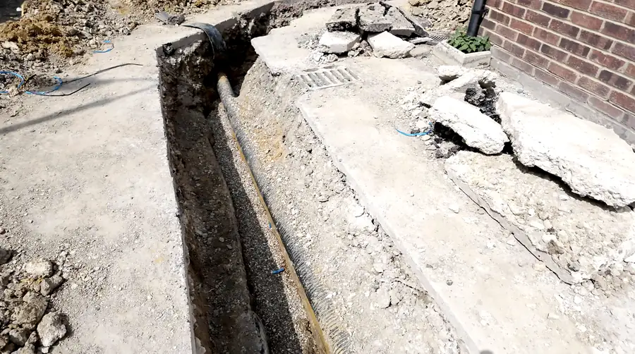 Narrow utility trench cut along a building wall with the duct visible